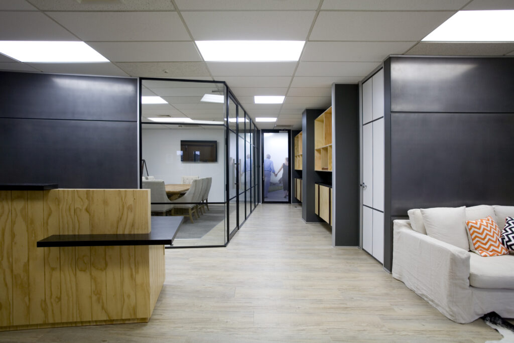 Modern office corridor with glass-partitioned meeting room, black and wood built-in cabinetry, light wood flooring, drop ceiling with LED panels, and a white sofa with patterned cushions - England