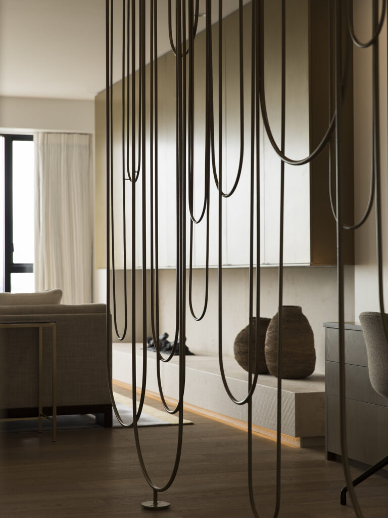 Contemporary living space featuring sculptural hanging bronze rod installation as a room divider, with brass cabinetry, stone bench seating, and warm timber flooring - Dubai