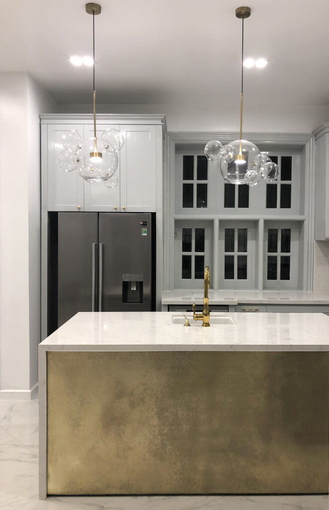 Contemporary kitchen interior featuring brass-clad island with patina finish, white stone benchtop, gold tapware, and glass pendant lights. Miami, Florida.