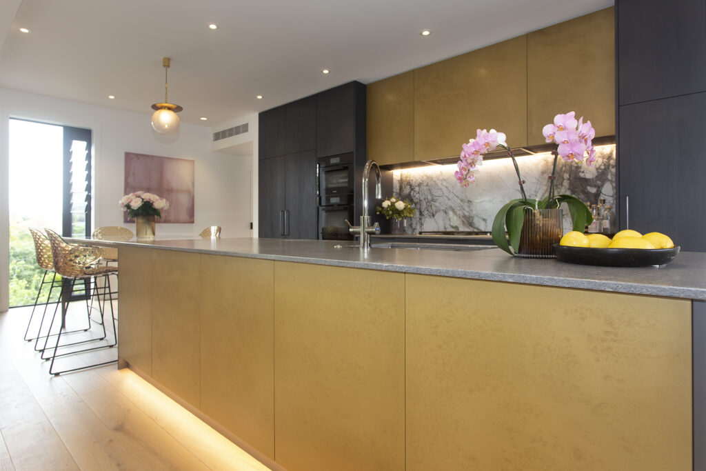 Contemporary kitchen with a large gold-toned island, gray countertop, marble backsplash, black cabinetry, pendant lighting, and bar stools, styled with orchids and a bowl of lemons - Sydney