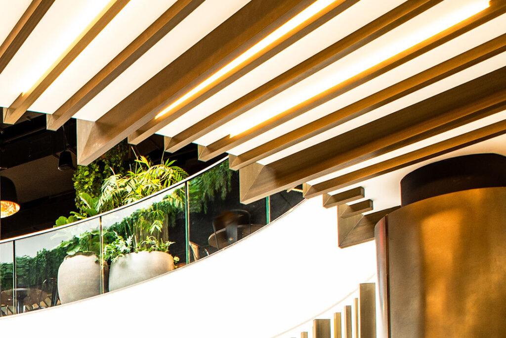 Curved shopping mall balcony with glass railing, large potted plants, and wood slatted ceiling with integrated lighting – Australia