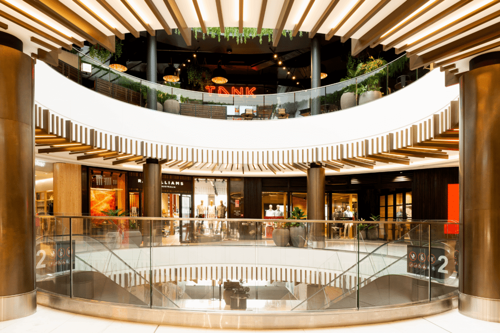 Modern circular shopping mall interior with curved balcony levels, wood slatted ceiling design, glass railings, and retail storefronts with ambient lighting – Australia